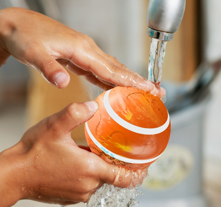 Orange automatic cat toy ball USB rechargeable being rinsed under running water from a faucet